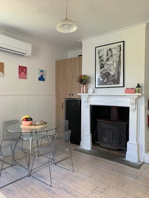 A dining area with a glass table and wire chairs, a black refrigerator, a wooden wardrobe, and a white fireplace with a framed picture above it.