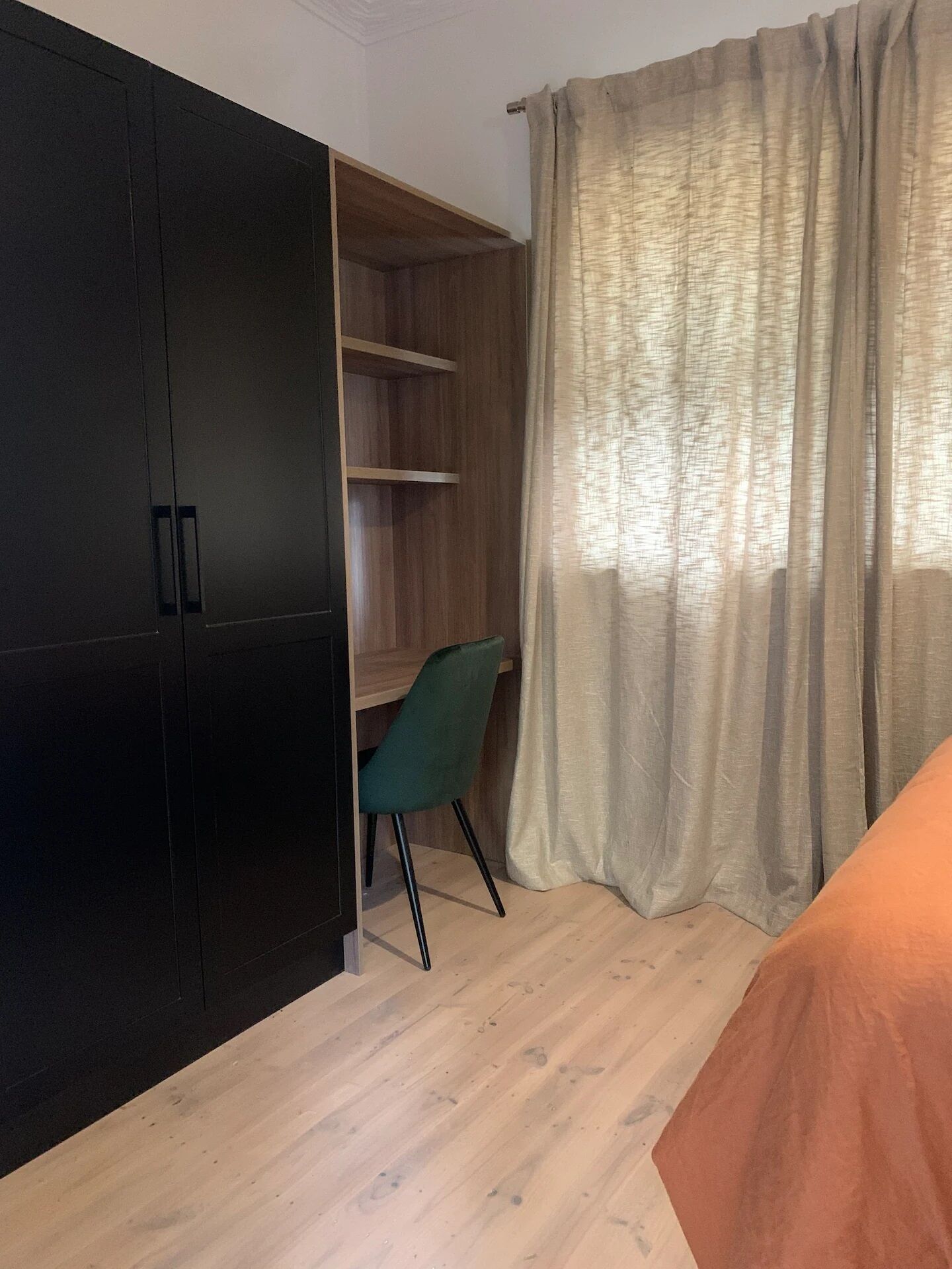 A room with a wooden wardrobe, a green chair, a bed with an orange blanket, and a window with beige curtains.
