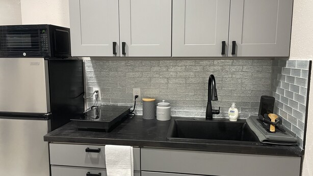 A compact kitchen with grey cabinets, a black countertop, a built-in oven, and a sink with a modern faucet. - Beautiful Studio in Venice, Fl (Venice)