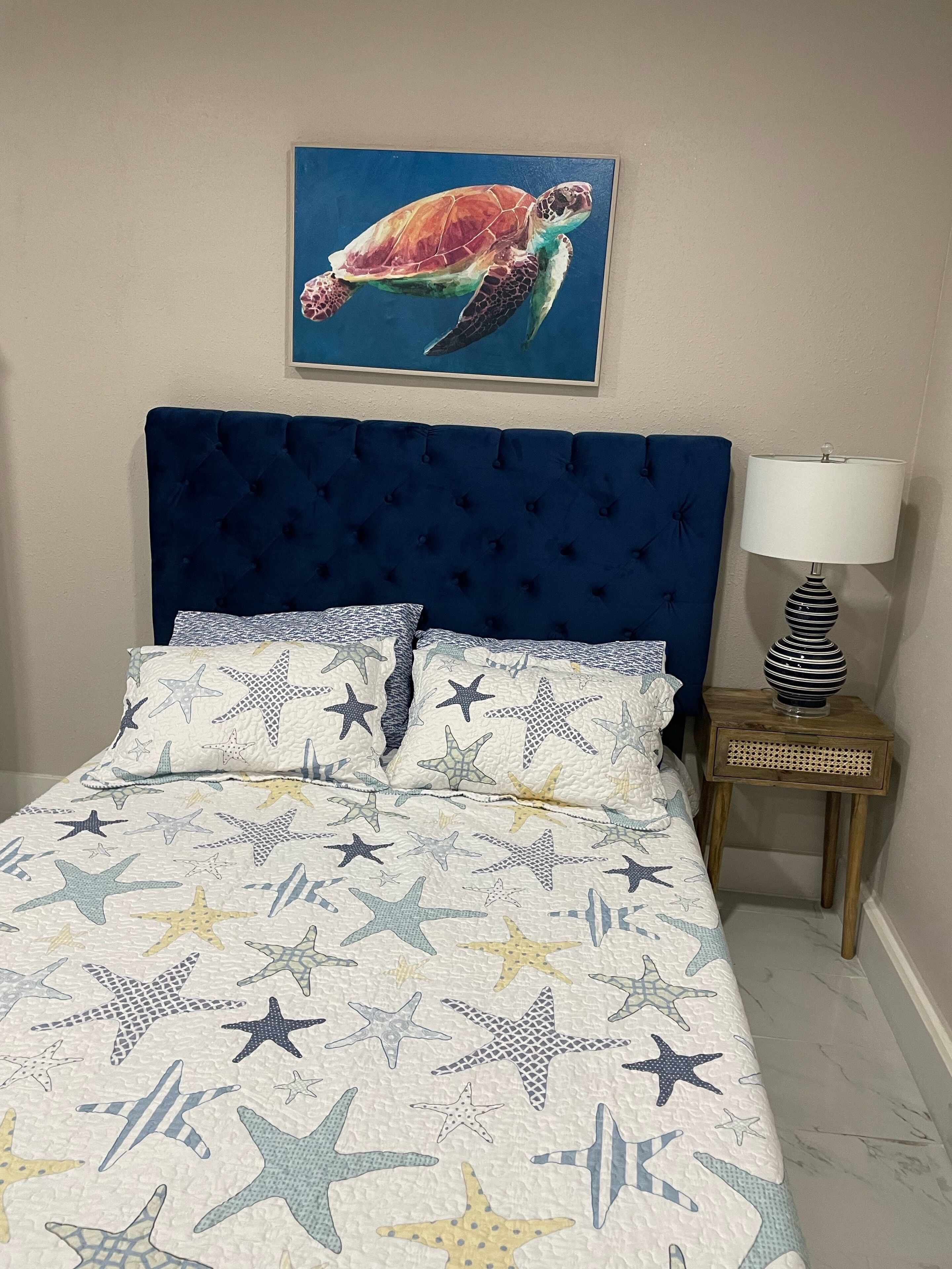 A bedroom with a blue headboard, star-patterned bedding, a framed sea turtle painting, and a bedside lamp.