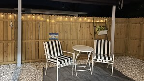 An outdoor patio with two striped chairs and a small table, a wooden fence, and string lights. - Beautiful Studio in Venice, Fl (Venice)