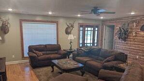Ein Wohnzimmer mit zwei braunen Ledersofas, einem Couchtisch und einem Hocker. An den Wänden befinden sich Hirschgeweihe und ein Deckenventilator.