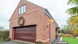 Un bâtiment en briques avec une porte de garage, une guirlande accrochée au mur et un jardin avec des plantes et un tonneau en bois.