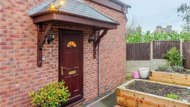 Une maison en briques avec une porte marron foncé, un petit porche et un jardin doté de plates-bandes surélevées.