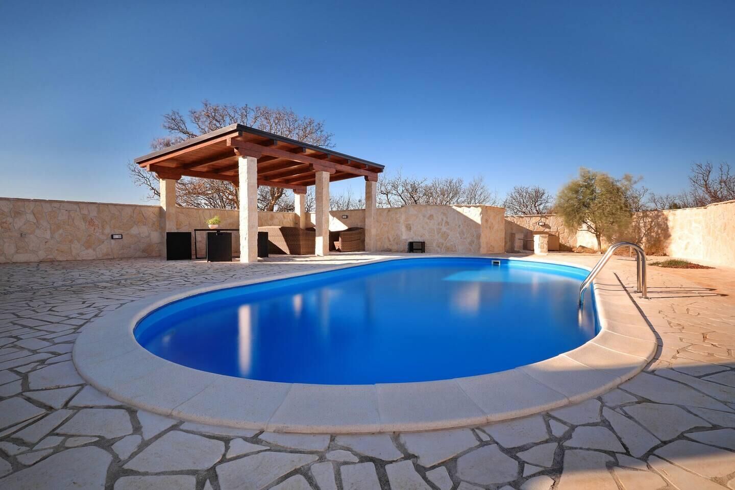 A swimming pool with a pergola structure, stone walls, and a clear blue sky.