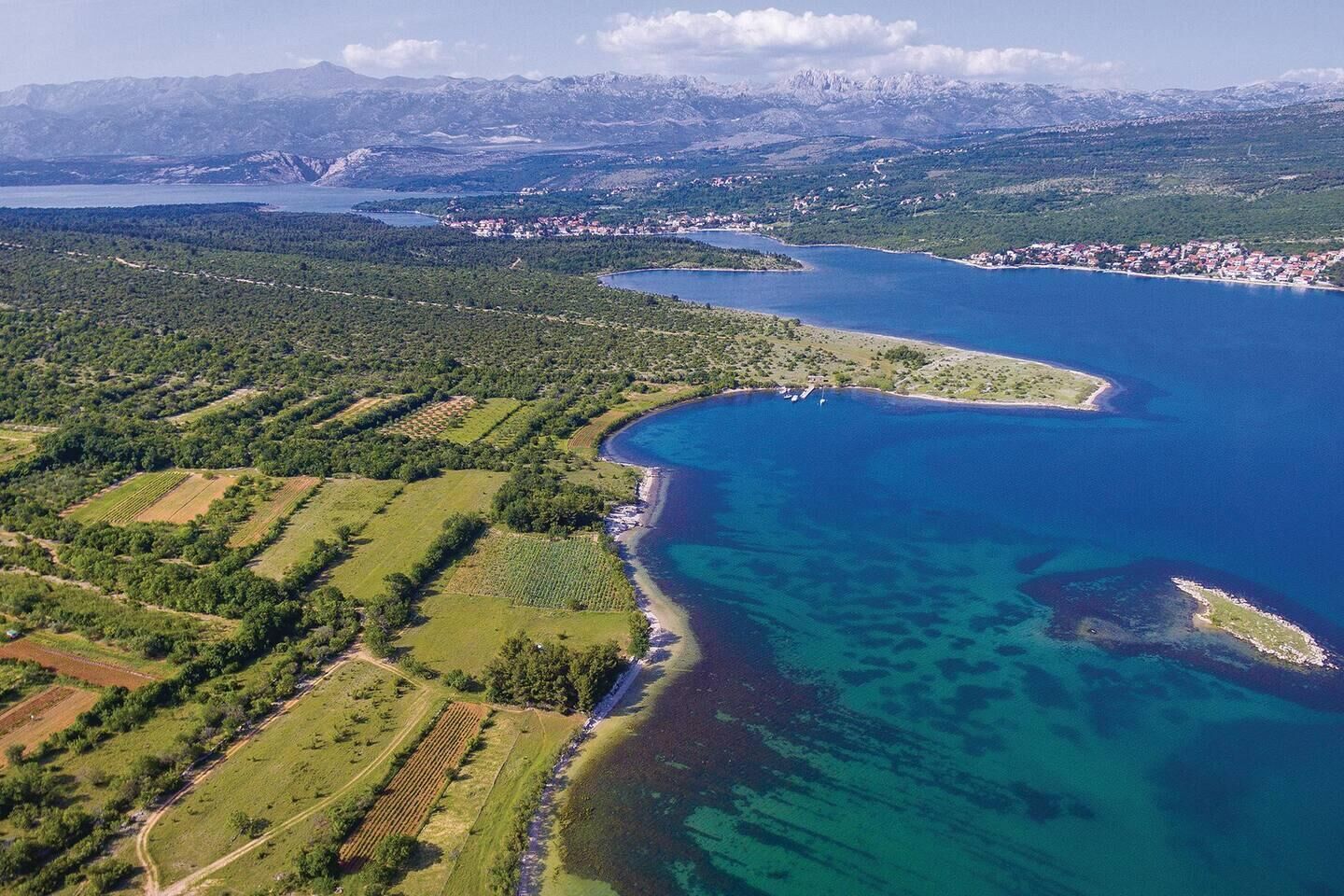 An aerial view of a coastal area with clear blue waters, green fields, and a small town in the background.