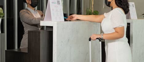 A hotel reception desk with a glass partition, a sign with health and safety guidelines, and a floral arrangement.