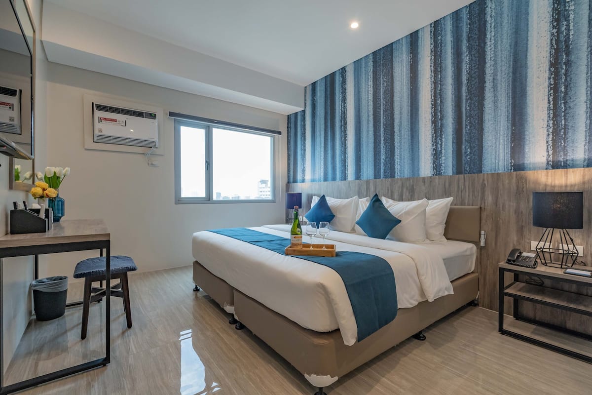 A modern hotel room with a large bed, a desk with a chair, a small table with a vase of flowers, a large window, and a wall-mounted air conditioner.