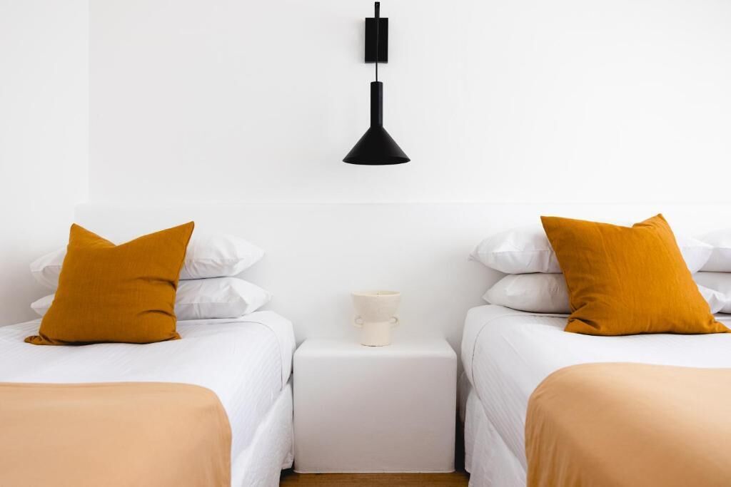 A hotel room with two beds, white bedding, mustard-colored pillows, a white nightstand with a white lamp, and a white cup.