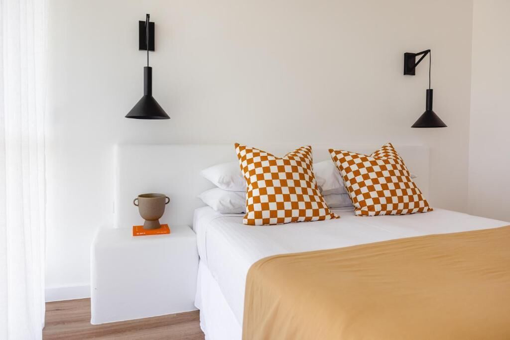 A neatly arranged hotel room with a bed, two checkered pillows, a bedside table with a cup, and two wall-mounted lamps.