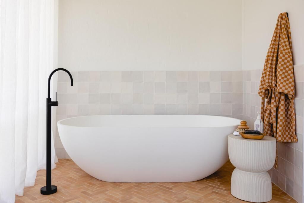 A modern bathroom with a freestanding bathtub, a towel rack with a checkered towel, and a small round stool with toiletries.