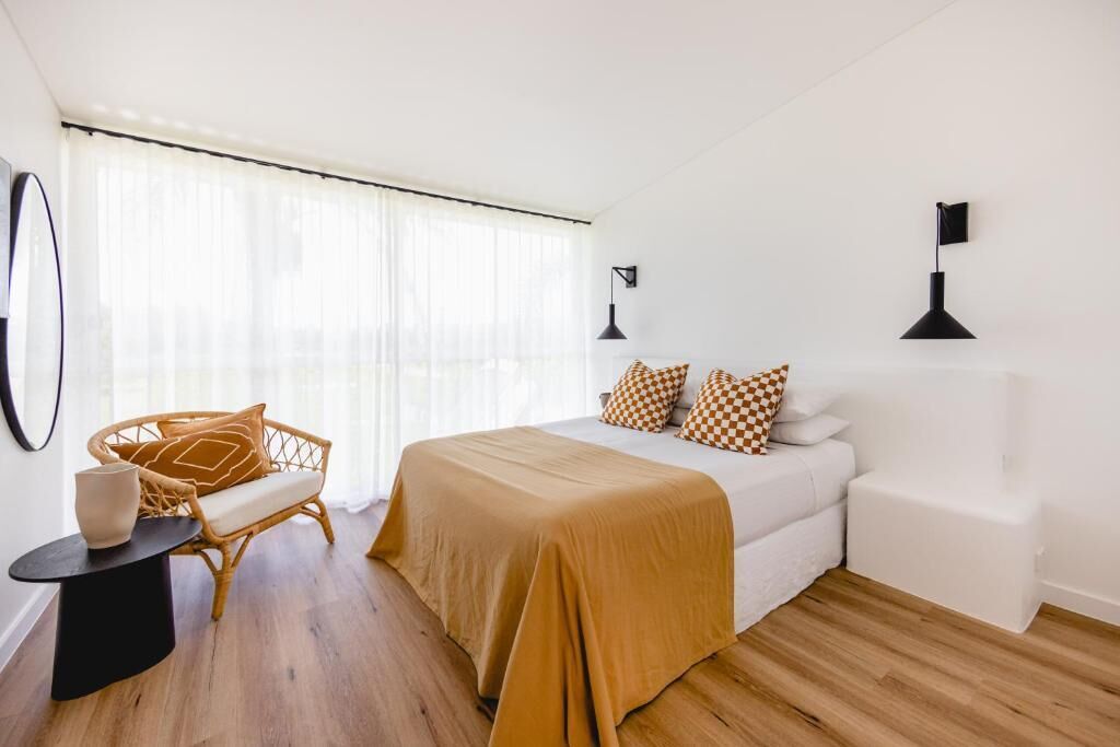 A bedroom with a bed, a chair, a small table, and a window with sheer curtains.