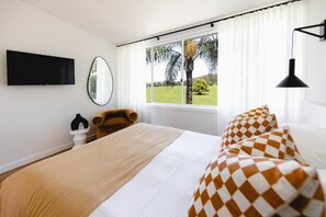 A hotel room with a bed, a television mounted on the wall, a chair, and a window with a view of a palm tree and greenery. - THE BERRY VIEW (Berry)