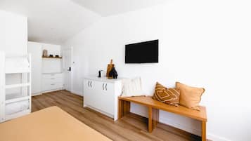 Un dormitorio con una cama litera, un banco de madera con almohadones, un aparador blanco y un televisor de pantalla plana montado en la pared.