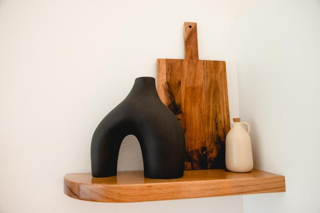 Una estantería de madera con un jarrón negro, una tabla de cortar de madera y una jarra blanca.