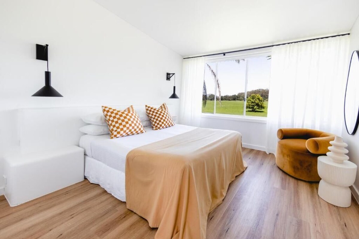 Ein modernes Schlafzimmer mit einem großen Bett, Kissen in Weiß und Orange, einer beigen Tagesdecke, einem weißen Sofa, einem senffarbenen Sessel und Blick auf eine grüne Landschaft durch große Fenster.