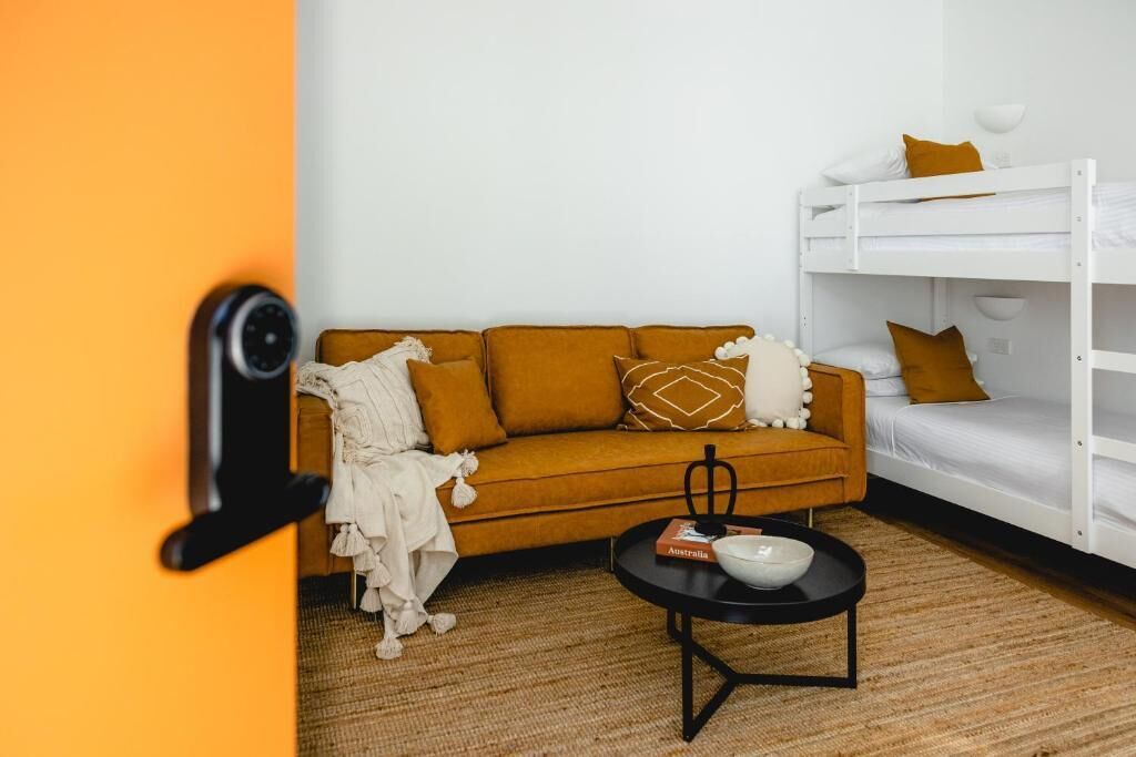 A room with a mustard-colored sofa, a white bunk bed, a black coffee table, and a door with a security camera.