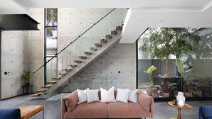 A modern living room with a staircase, a sofa with cushions, a coffee table with books and a vase, and a potted plant.