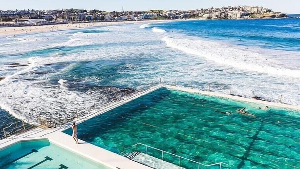 A pool adjacent to a beach with clear blue water, a rocky shoreline, and a town in the background.