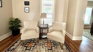 Una sala de estar con dos sillones blancos, un tapete estampado, una mesita con lámpara y un ventanal con persianas.