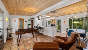 Ampla sala de estar com mesa de futebol, poltronas de couro e cozinha ao fundo.