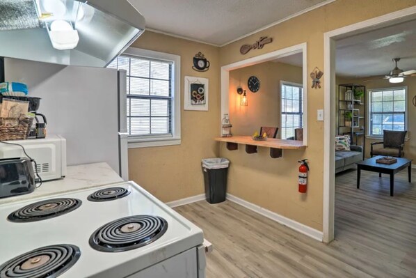 Cocina compacta con estufa, microondas y un pequeño cubo de basura. Se ve una ventana con persianas, un estante en la pared con objetos decorativos y un extintor.