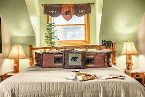 A bedroom with a bed, two bedside lamps, a window with a view, a small Christmas tree, and a bear-themed pillowcase.