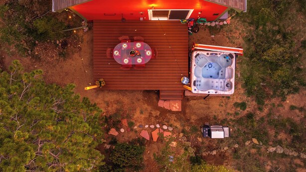 A hot tub and a table with chairs on a wooden deck, surrounded by rocks and trees.