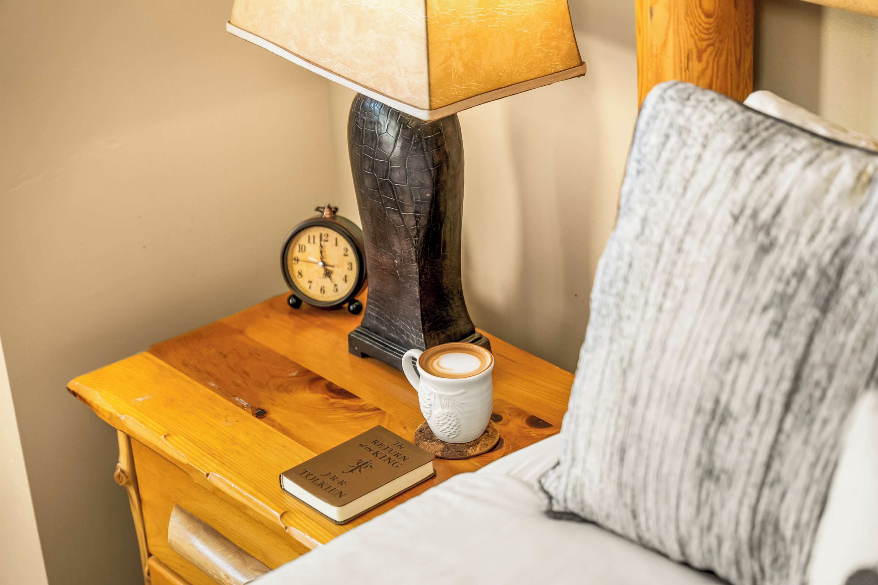 A wooden bedside table with a lamp, a book titled 'The Return of the King' by J.R.R. Tolkien, a cup of coffee, and a pillow.