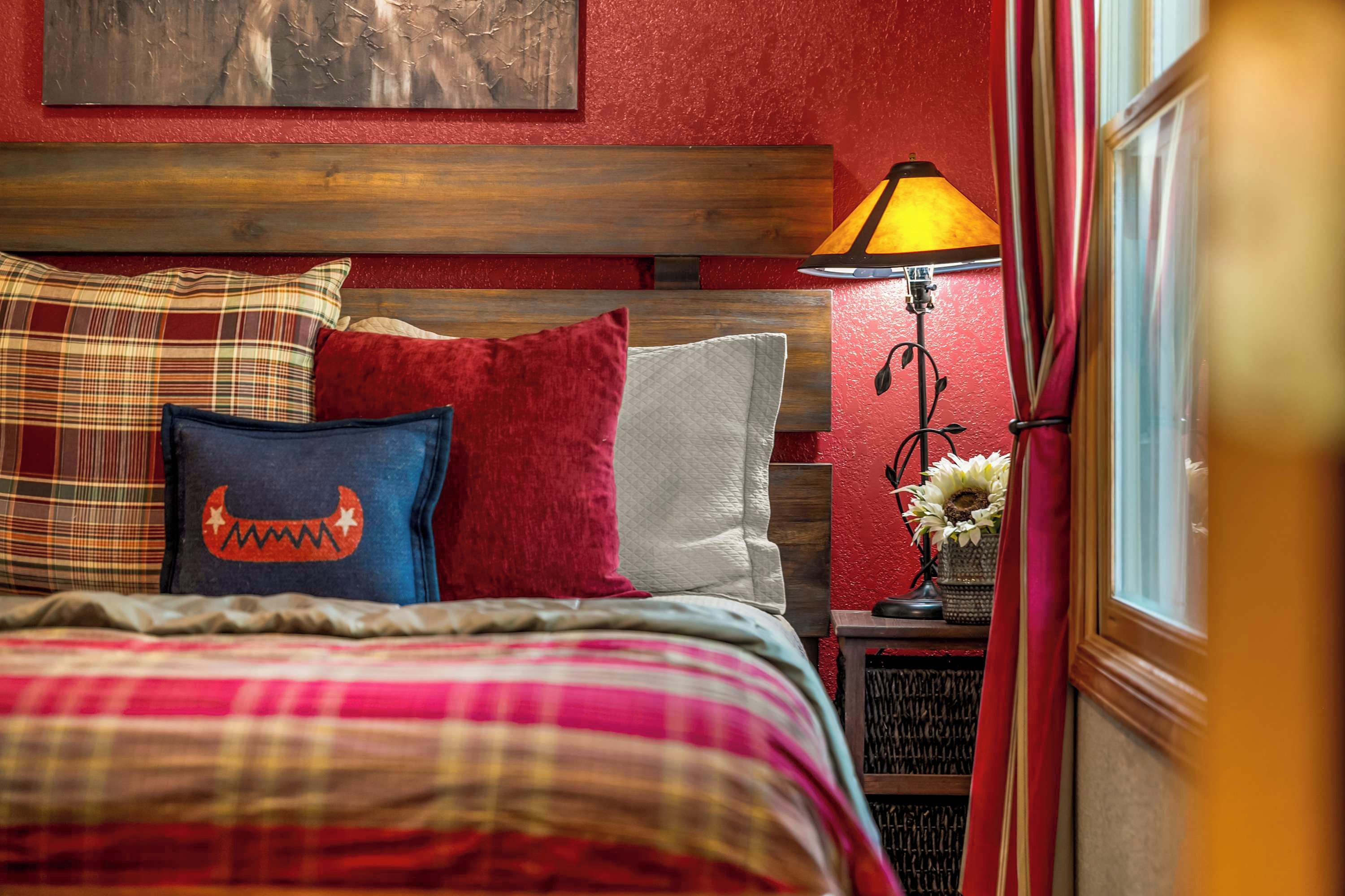 A bed with a plaid bedspread and multiple pillows, a nightstand with a lamp and a vase of flowers, and a red textured wall.