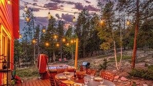 An outdoor dining area with string lights, a hot tub, and a deck overlooking a forested area.