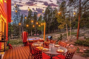 An outdoor dining area with string lights, a hot tub, and a deck overlooking a forested area.