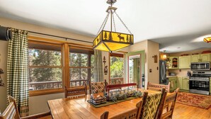 A dining area with a wooden table and chairs, a hanging light fixture, and a view of the outdoors through large windows.
