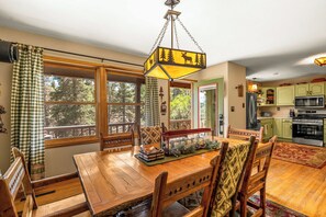 A dining area with a wooden table and chairs, a hanging light fixture, and a view of the outdoors through large windows.