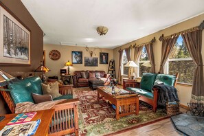 A cozy living room with a green sofa, a wooden coffee table, and a patterned rug. There are framed pictures on the walls and a window with curtains.