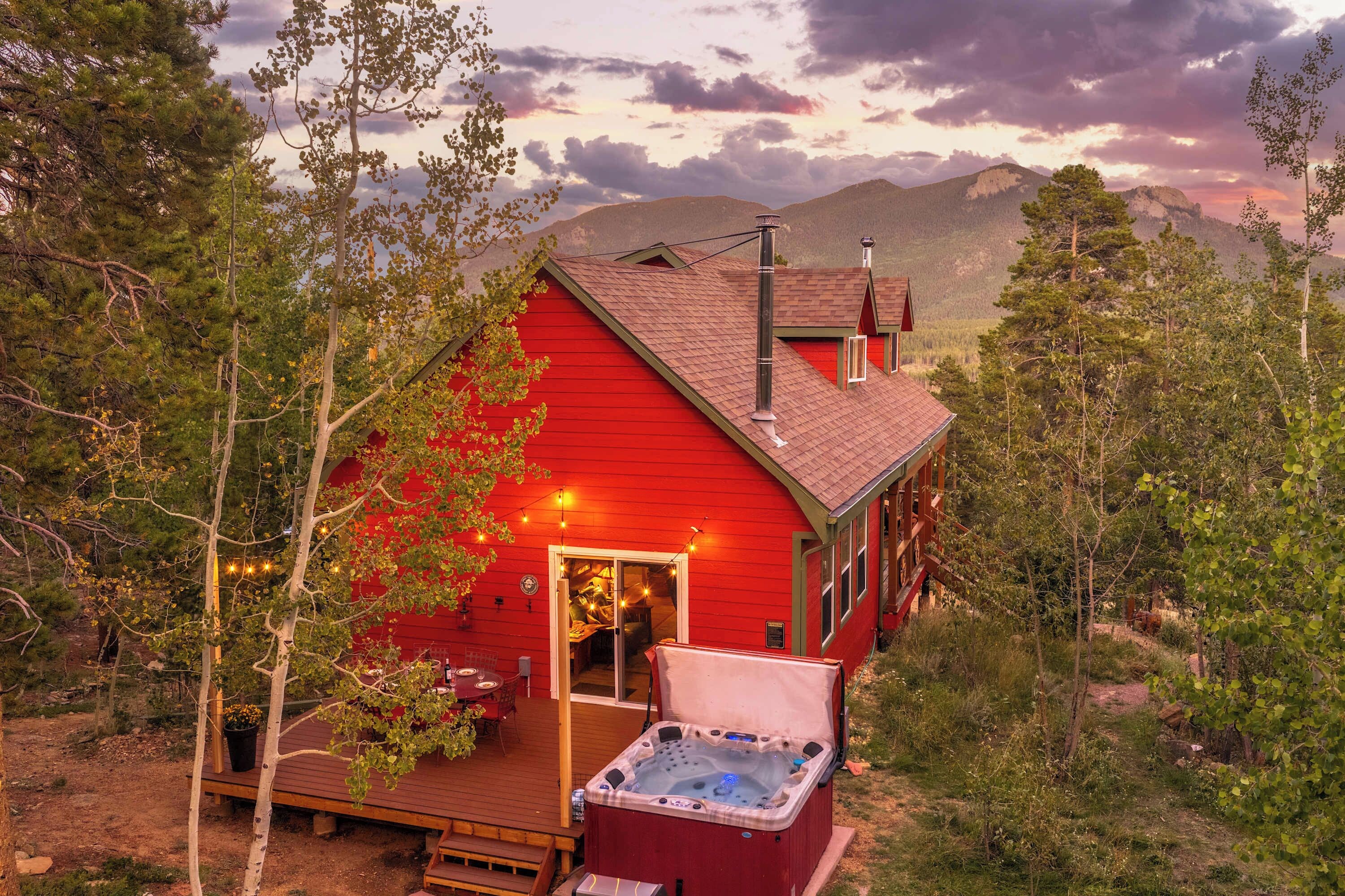 Eine rote Holzhütte mit Whirlpool und einer Terrasse, umgeben von Bäumen und Bergen.