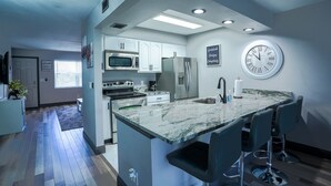 Una cocina moderna con armarios blancos, horno empotrado, microondas y refrigerador. Hay una isla grande con taburetes de bar y un reloj montado en la pared.