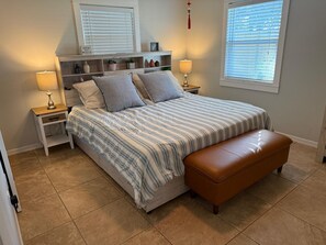 A bedroom with a bed, bedside tables, a bench, a shelf with decorative items, and a window with blinds.