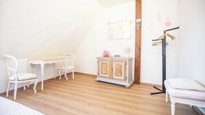 Quarto com teto inclinado, piso de madeira, toucador branco com espelho, mesa pequena, duas cadeiras brancas, armário e cama com travesseiro branco.