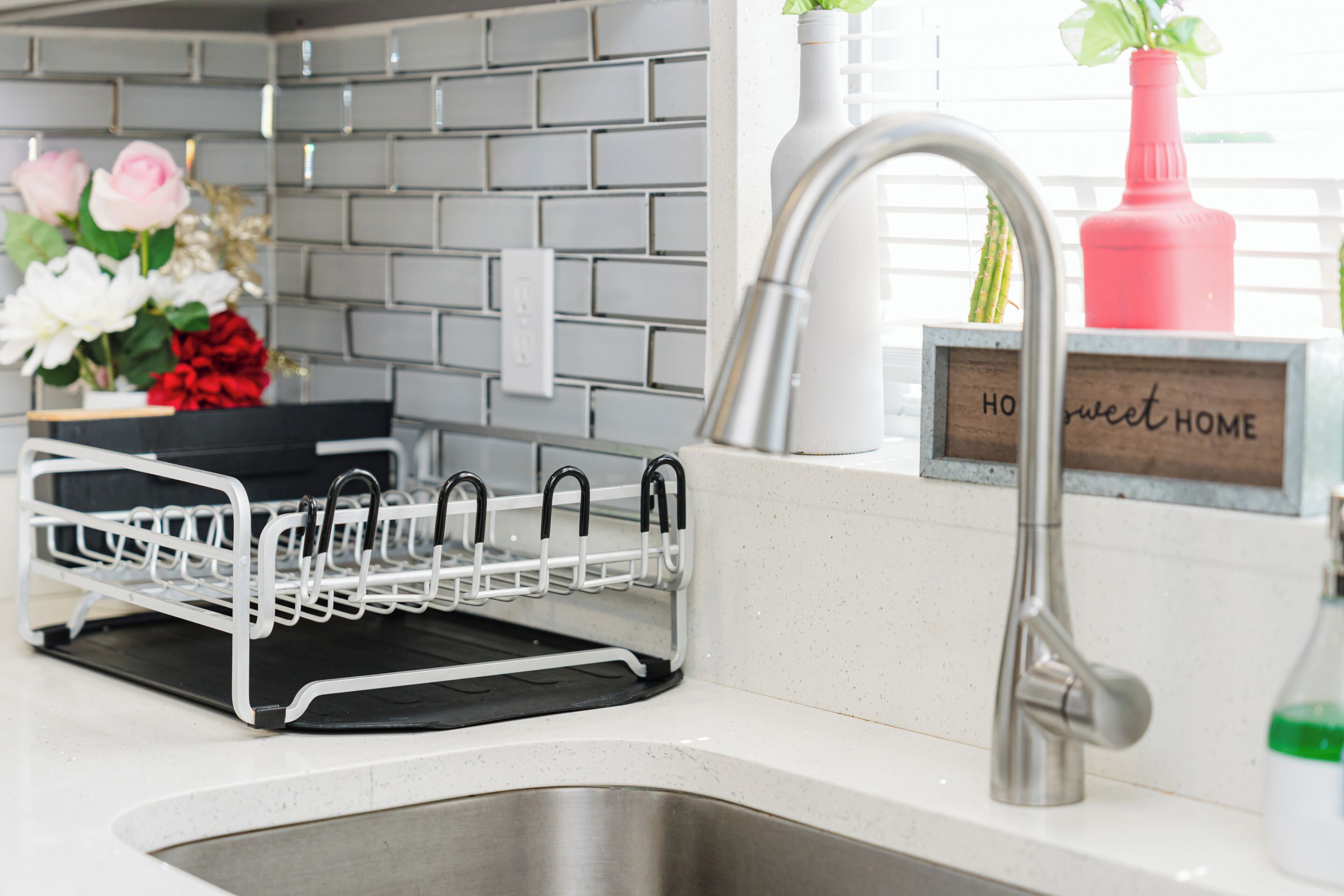 Una cocina con un lavamanos de acero inoxidable, un escurreplatos, una planta en maceta y un letrero que dice 'HOw sweet HOME'.