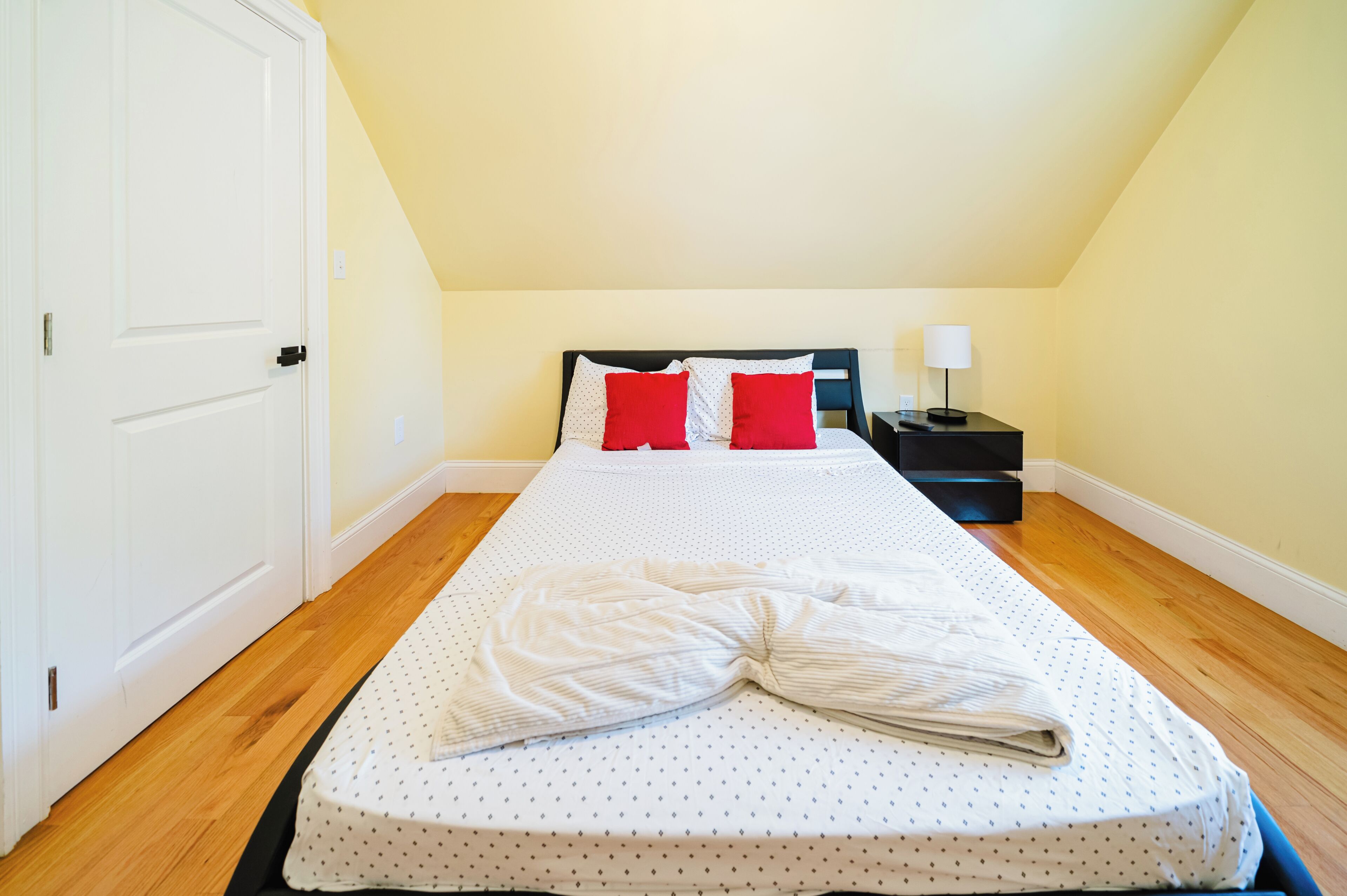 Una cama individual con colchón blanco y almohadas rojas, una mesita de noche con lámpara, y una puerta a la izquierda.