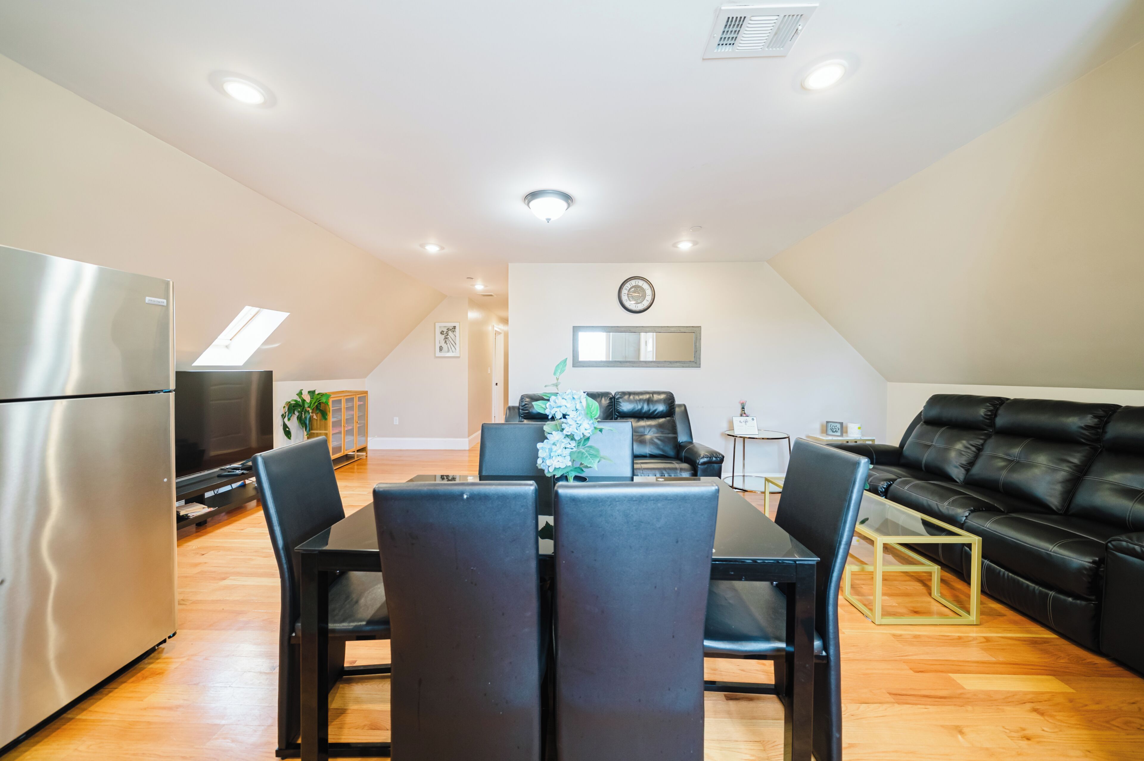 Una sala de estar moderna con un sofá de cuero negro, una mesa de comedor con sillas, un televisor y un refrigerador.