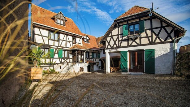 A traditional half-timbered building with a gravel courtyard, wooden benches, and a staircase leading to the entrance. - Gîtes Rocher des Celtes (Dieffenthal)