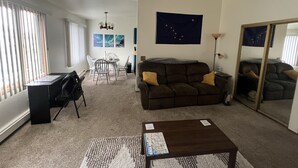 A living room with a brown sofa, a coffee table, a dining table with chairs, and a large window with blinds.