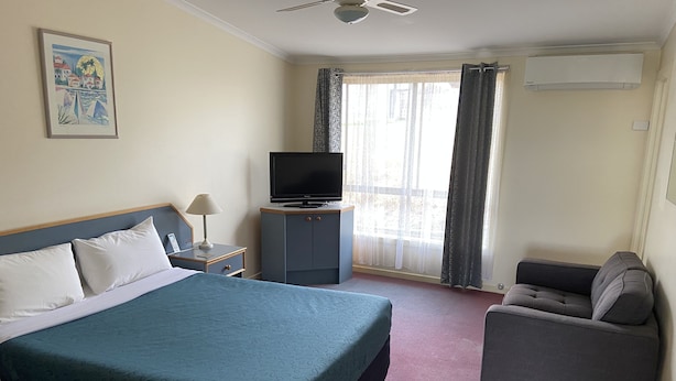 Une chambre d’hôtel avec un lit, un téléviseur posé sur un meuble, un bureau avec une lampe, une chaise et une fenêtre avec des rideaux.