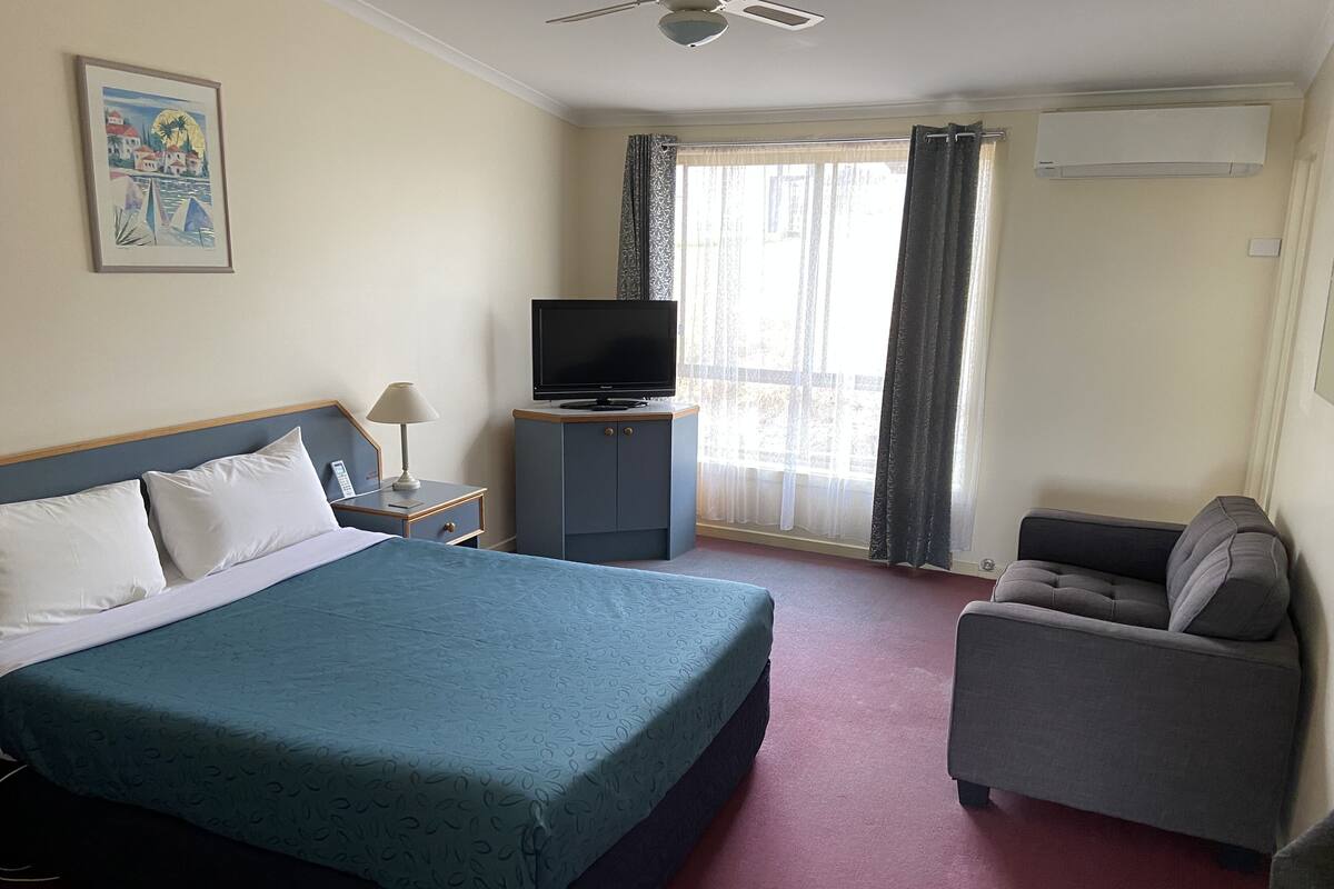 A hotel room with a bed, a television on a stand, a desk with a lamp, a chair, and a window with curtains.