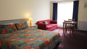 Une chambre d’hôtel avec un lit, un canapé rouge, une table en bois avec des chaises, une lampe et une fenêtre avec des rideaux.