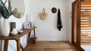 A hallway with a wooden floor, a white door, a mirror, a table with decorative items, and a wooden door with a view of greenery. - Deja Blue Anna Bay (Anna Bay)