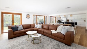 A spacious living area with a brown leather sectional sofa, two round coffee tables, and a large window providing natural light. - Deja Blue Anna Bay (Anna Bay)