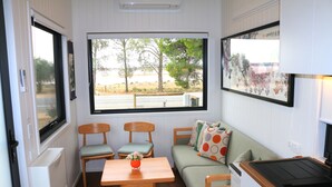 Interior compacto de una autocaravana con zona de comedor, asientos y un amplio ventanal que ofrece vistas a árboles y un cuerpo de agua.